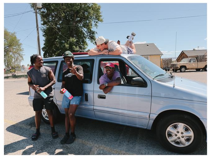 Four friends laughing around a light blue minivan, one lying on the roof