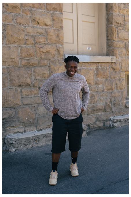 Smiling man in a camo hoodie and black shorts in front of a stone building