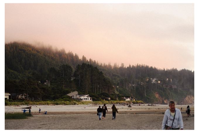 People walking along a beach toward a misty forested hillside at pink sunset