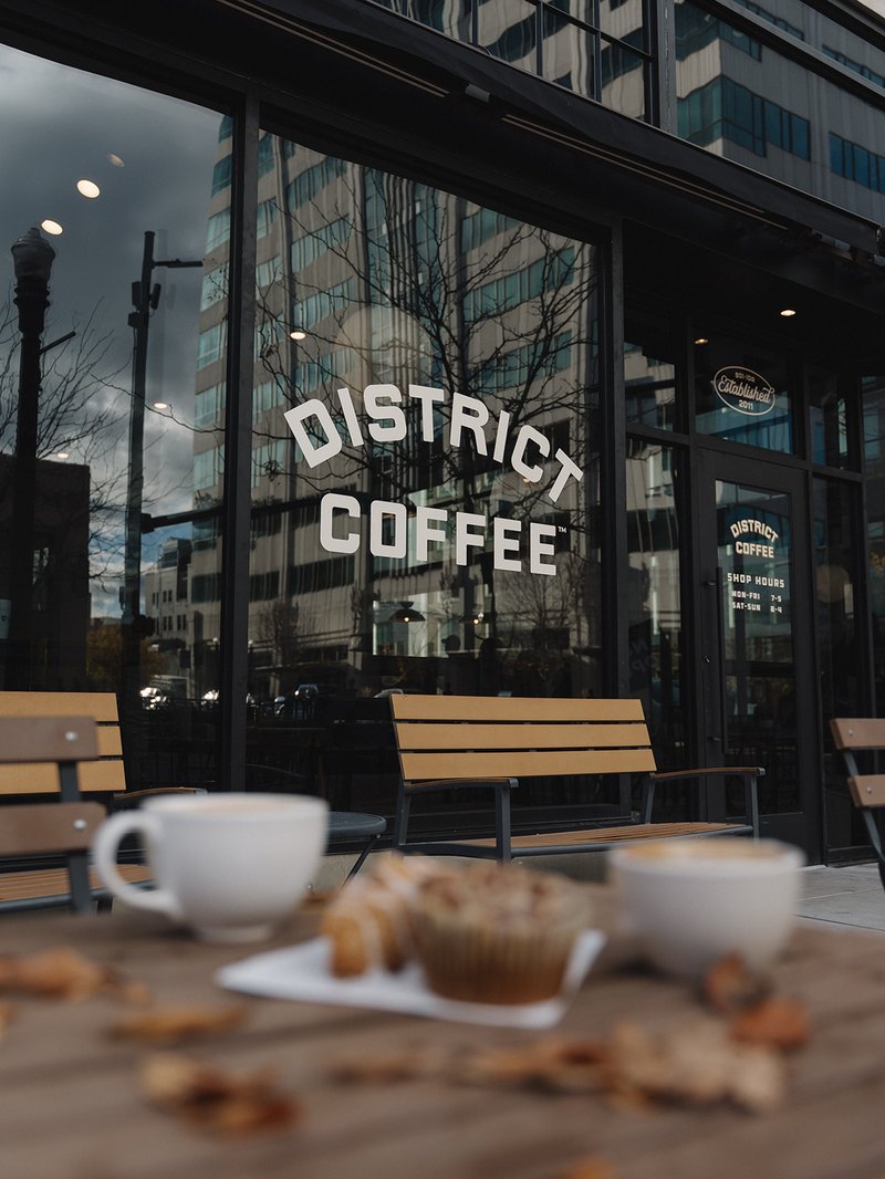 District Coffee window lettering reflecting downtown buildings, with two blurred lattes and a pastry on a patio table in the foreground