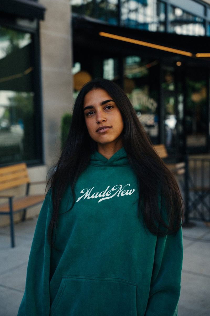 A woman with long dark hair looking at the camera in a green Made New hoodie on a city sidewalk outside a cafe