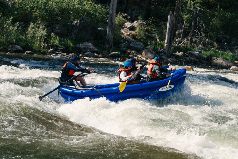 A blue RIVTEK raft with a guide on oars and three paddlers in helmets pushing through a churning whitewater rapid