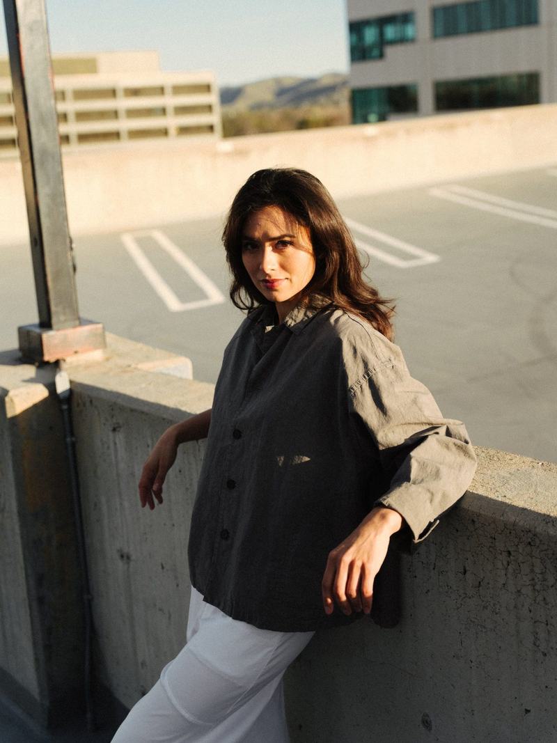 A woman in a gray linen jacket and white trousers leaning on a rooftop wall in golden light, looking toward the camera