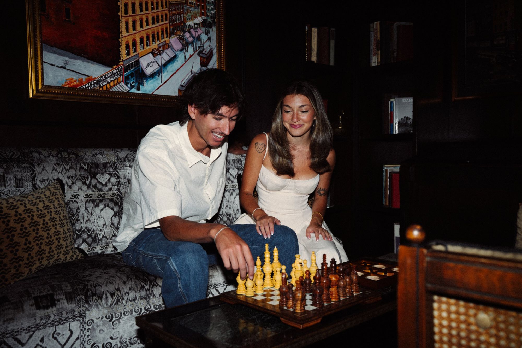 Lucas and Audry laughing over a chess game on a patterned sofa in a wood-paneled room