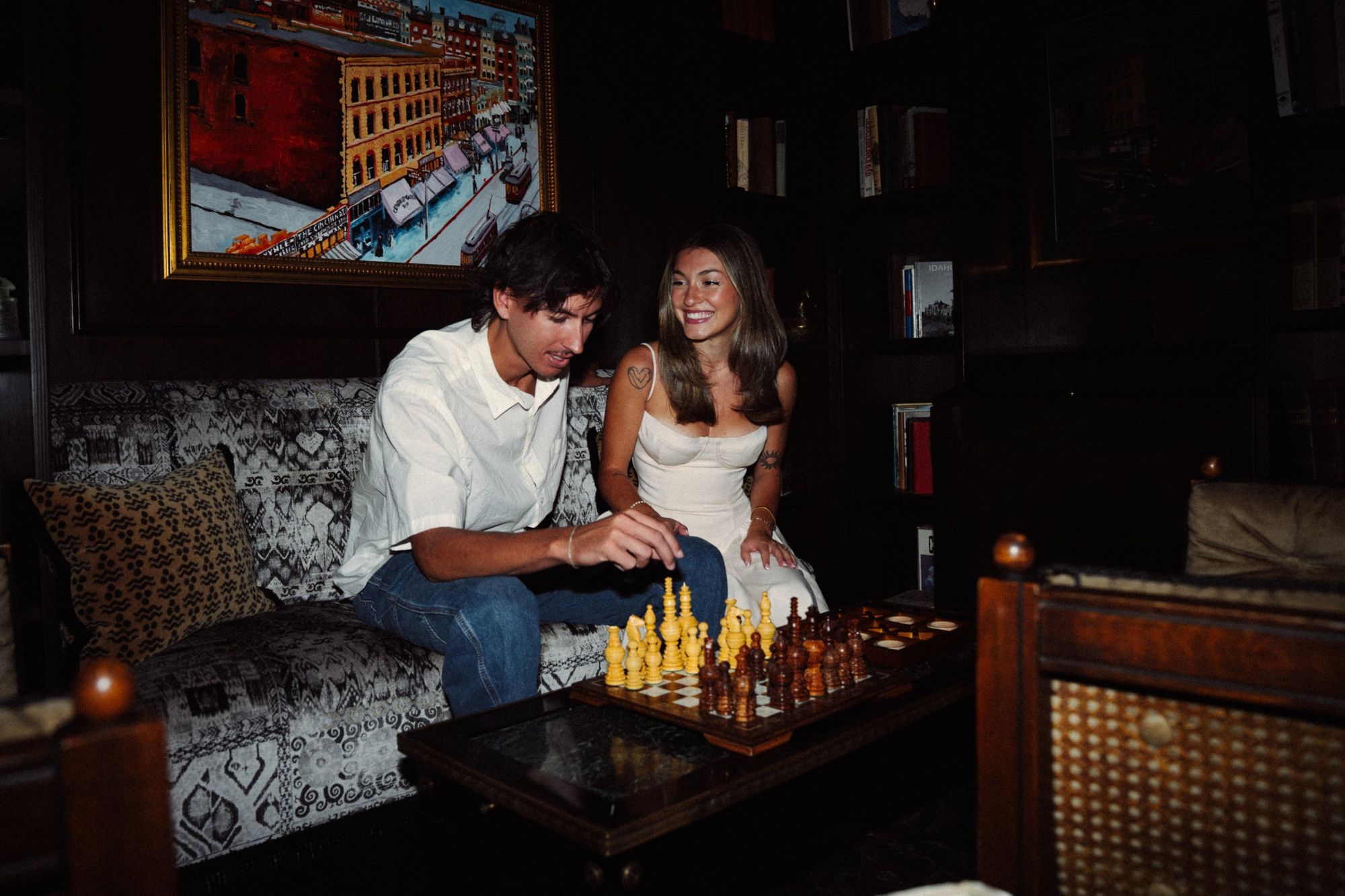 Audry laughing as Lucas reaches for a chess piece, both seated on a patterned sofa