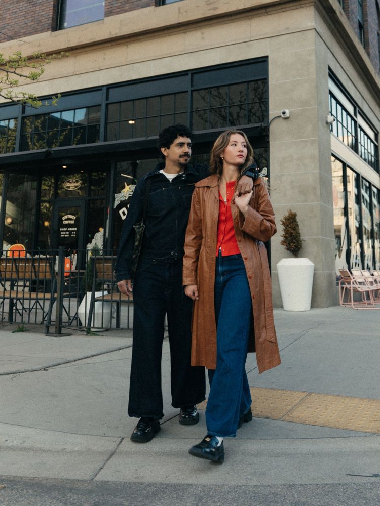 Aubrey and her partner walking arm in arm past a downtown Boise coffee shop, she in a brown leather trench and red cardigan, he in dark denim