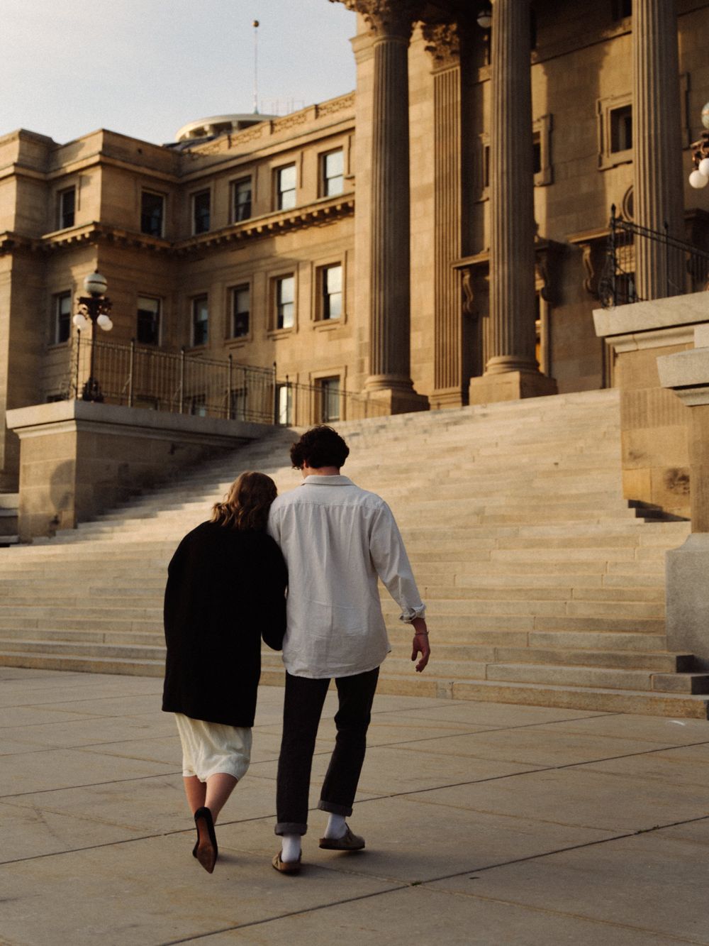 A couple walking away from the camera toward the Idaho State Capitol steps at golden hour, she leaning her head on his shoulder