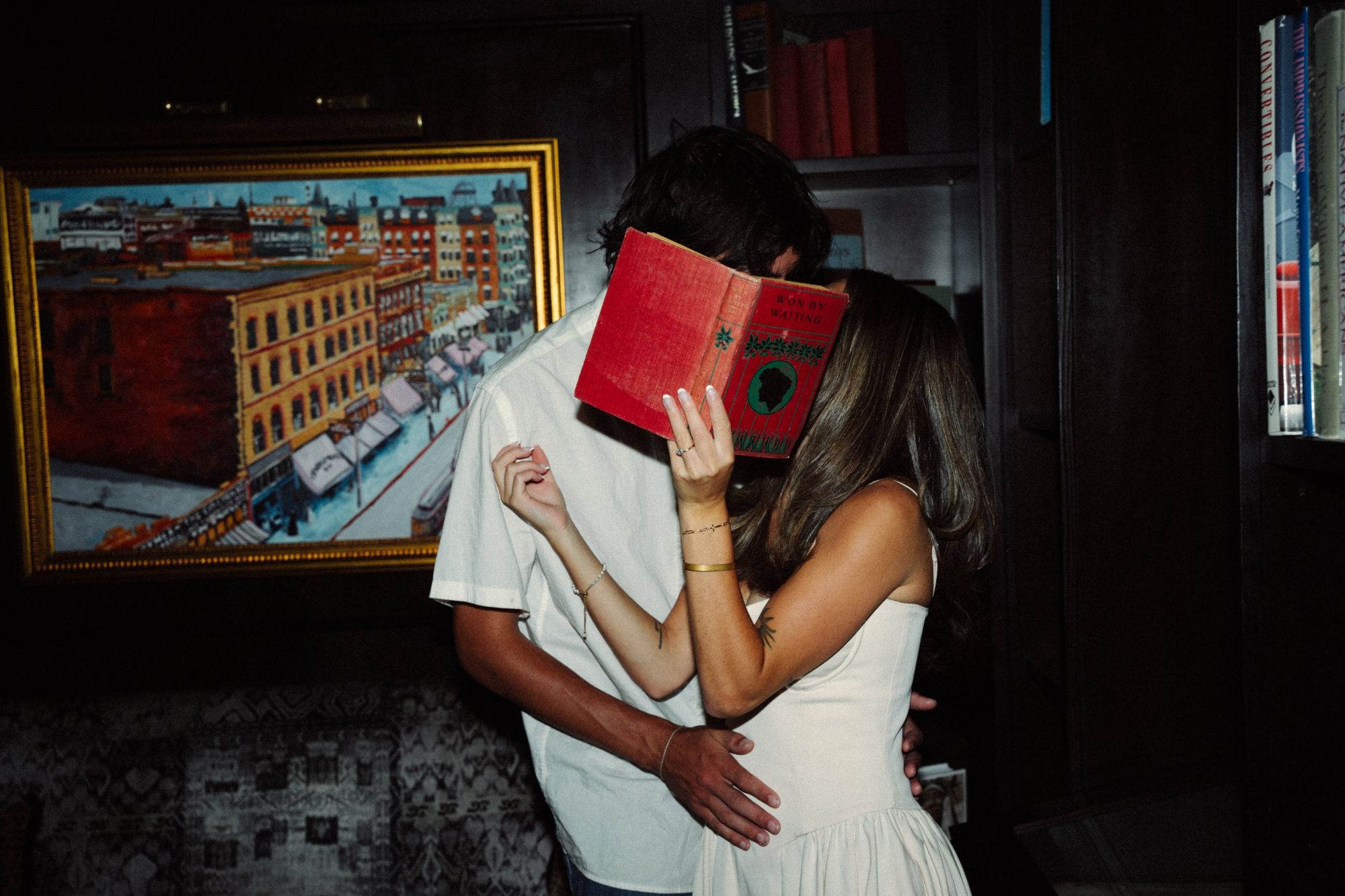 Lucas and Audry kissing behind a red vintage book in front of a painting of a city street