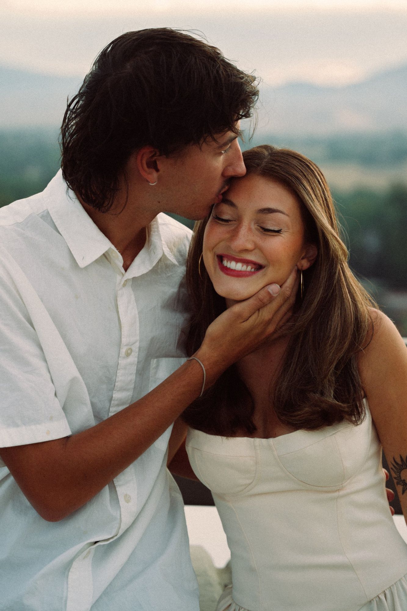 Close-up of Lucas kissing Audry's forehead as she laughs with her eyes closed, soft hills behind them