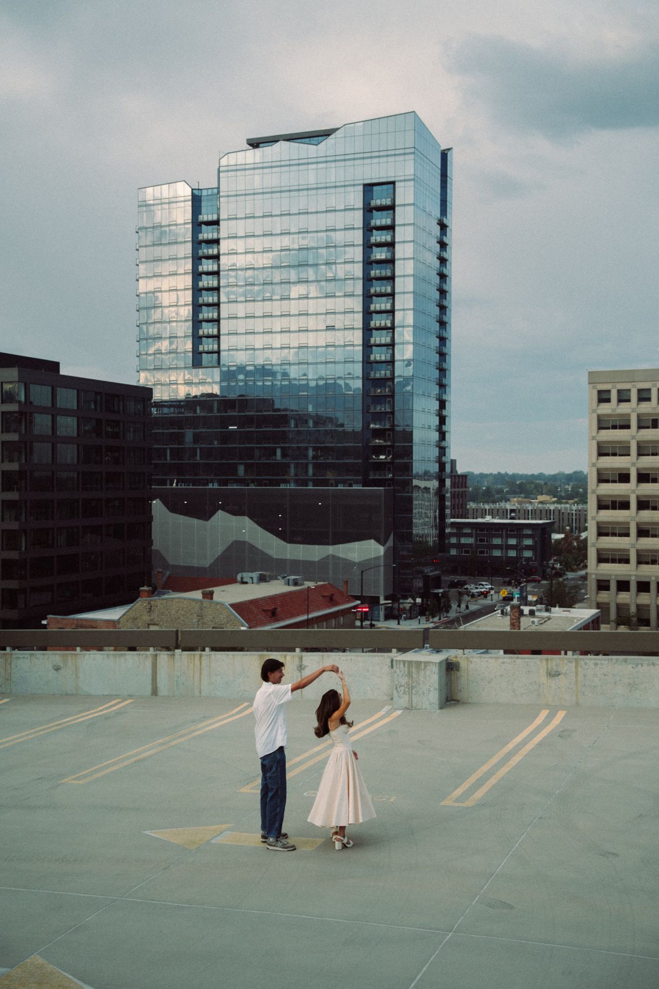 Lucas twirls Audry on a rooftop parking deck in front of a tall glass building in downtown Boise