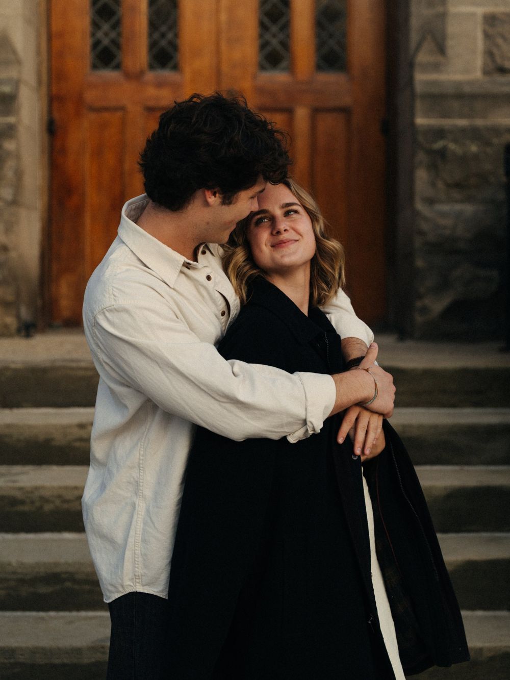 A man in a cream shirt hugging his partner from behind in front of a wooden church door while she looks up at him and smiles