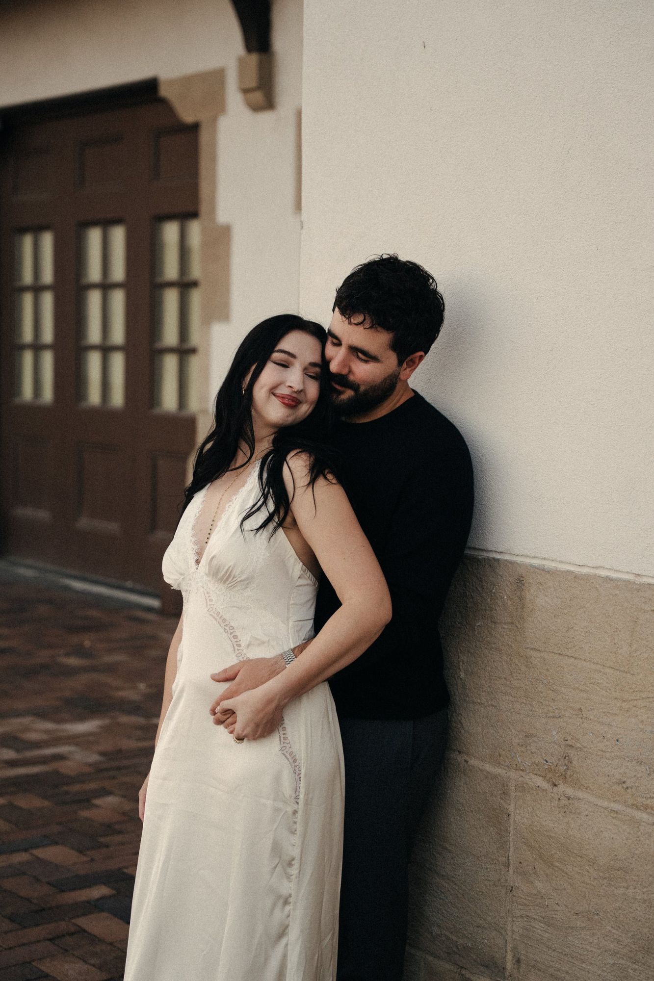James holds Cora from behind against a sandstone and stucco wall at the Boise Depot as she smiles with her eyes closed
