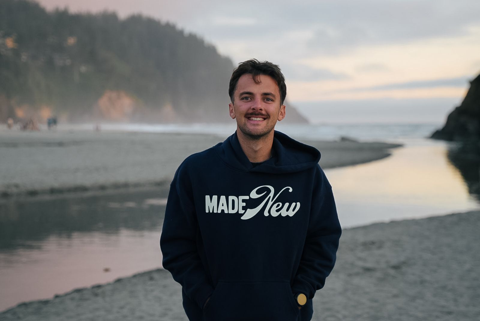 Trace Keller, a Boise brand photographer and videographer, smiling in a Made New hoodie on a beach at sunset