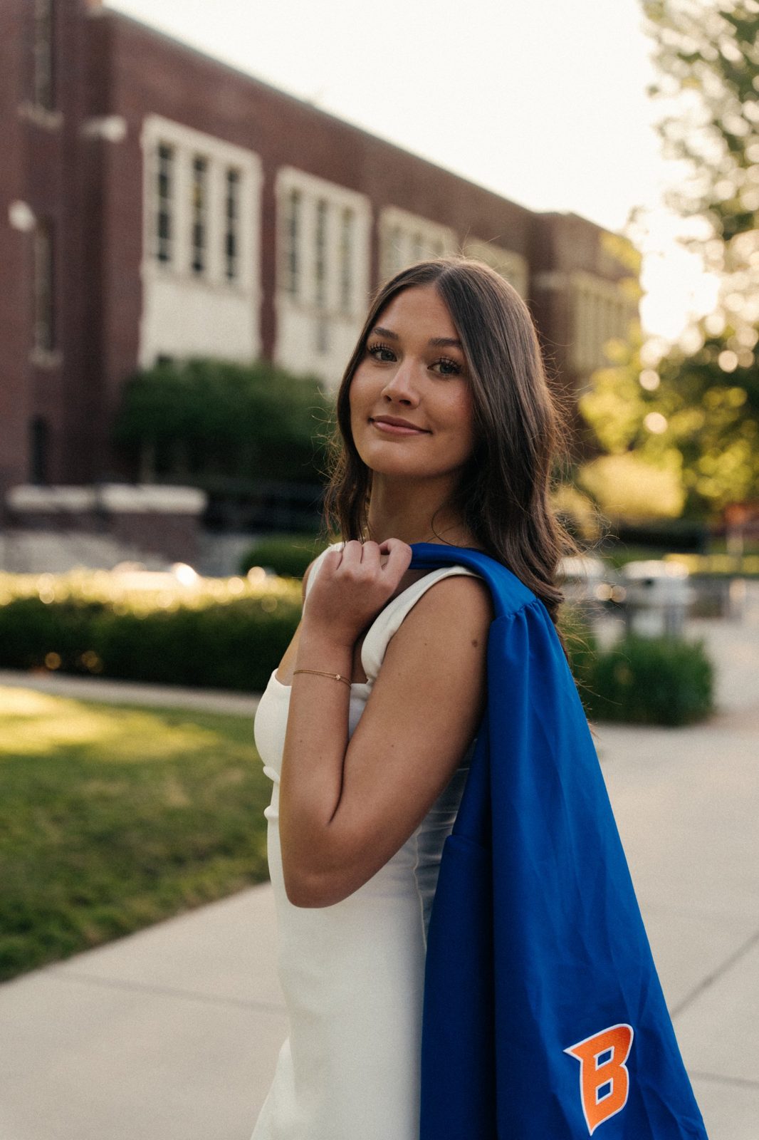Charlotte glancing back over her shoulder with a blue Boise State graduation gown draped on it, on a sunlit campus walkway