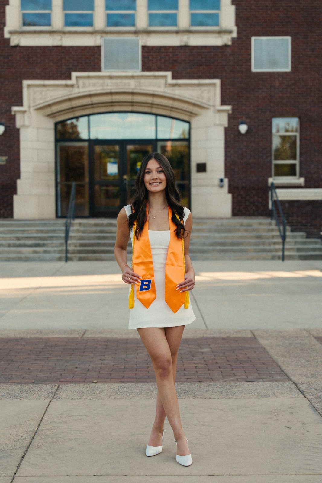 Charlotte in a white dress holding her orange Boise State graduation stole, standing in front of a brick building with a smile