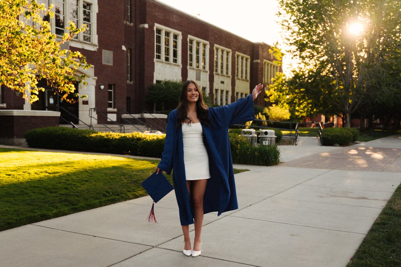 Charlotte in her blue gown holding her cap and waving on a sunlit Boise State campus walkway