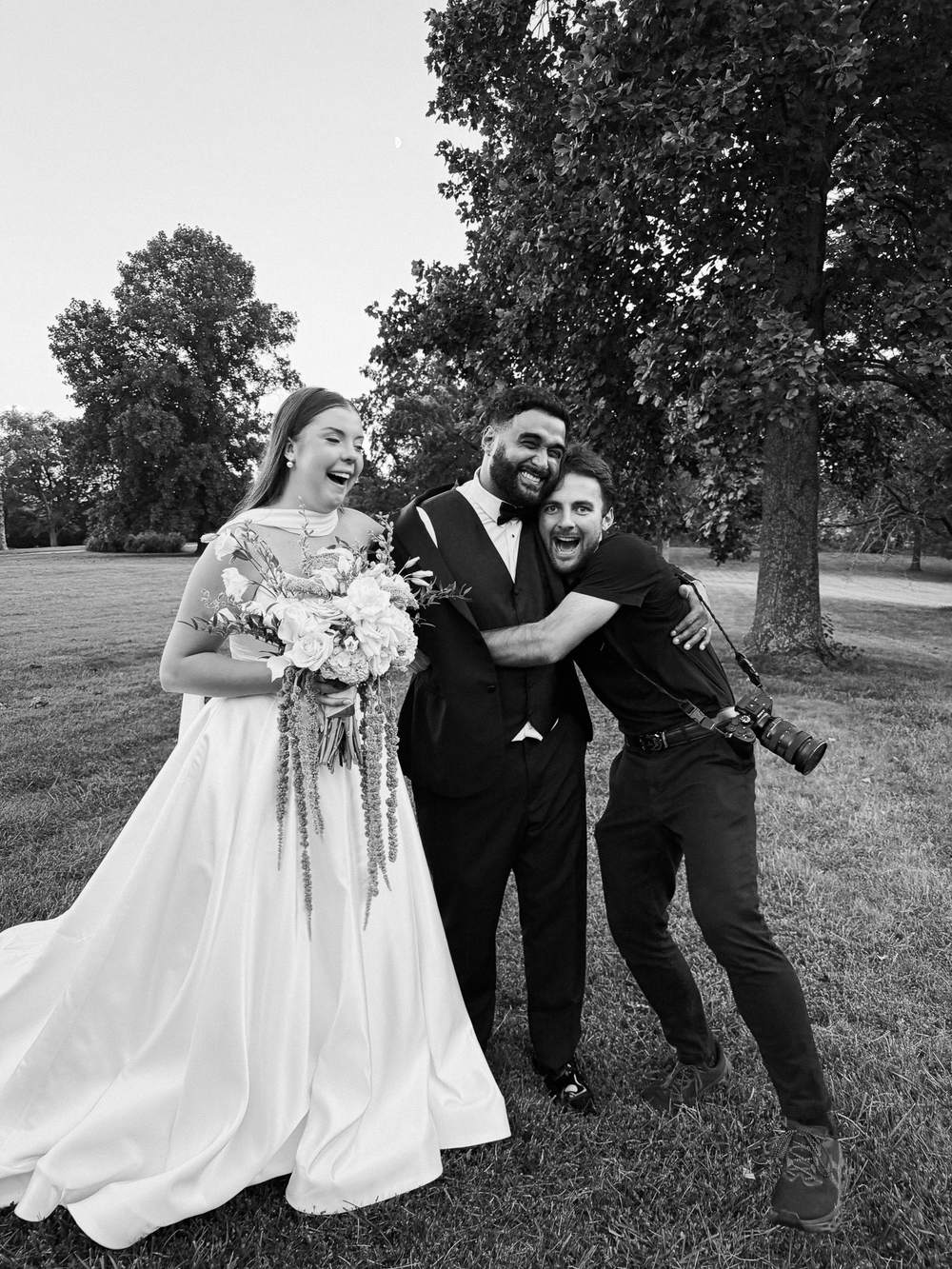 A bride and groom laughing as they hug their photographer in a park, black and white