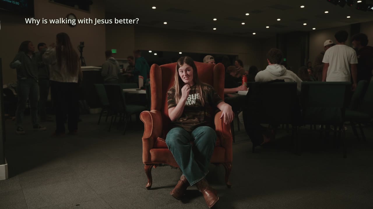A student sitting in an orange armchair answering a question in a dim room, with other students gathered behind her