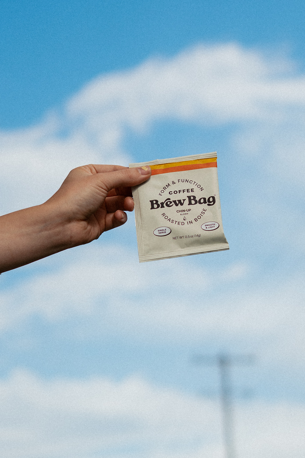 Hand holding up a Brew Bag single-serve coffee packet against a bright blue sky with clouds