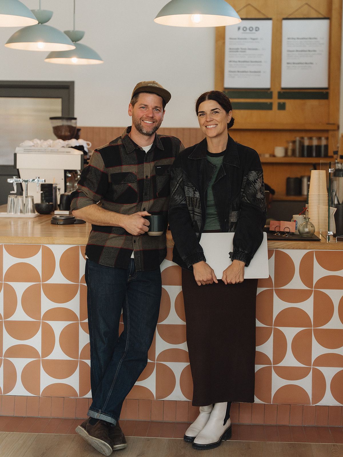 Man in a flannel shirt holding a mug and woman holding a laptop, smiling as they lean against the tiled counter at the District Coffee House