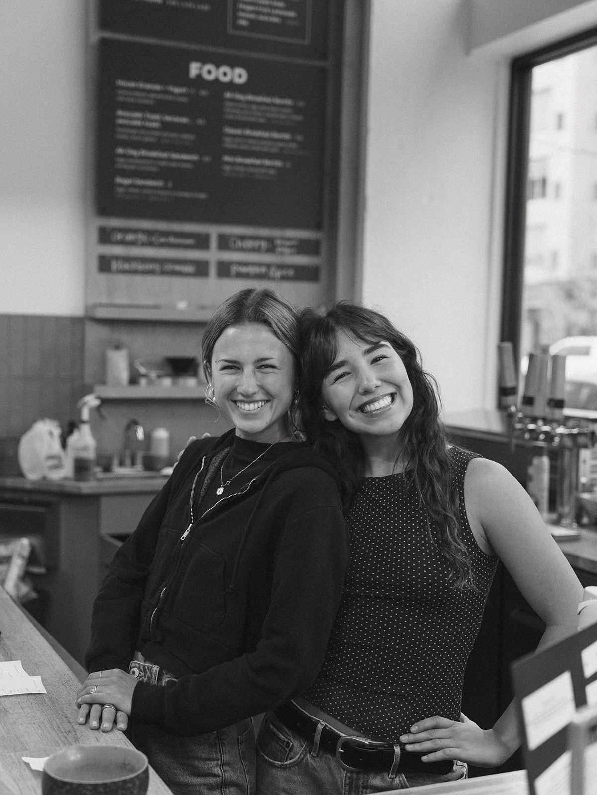 Black and white photo of two baristas laughing with their heads tipped together in front of the food menu board