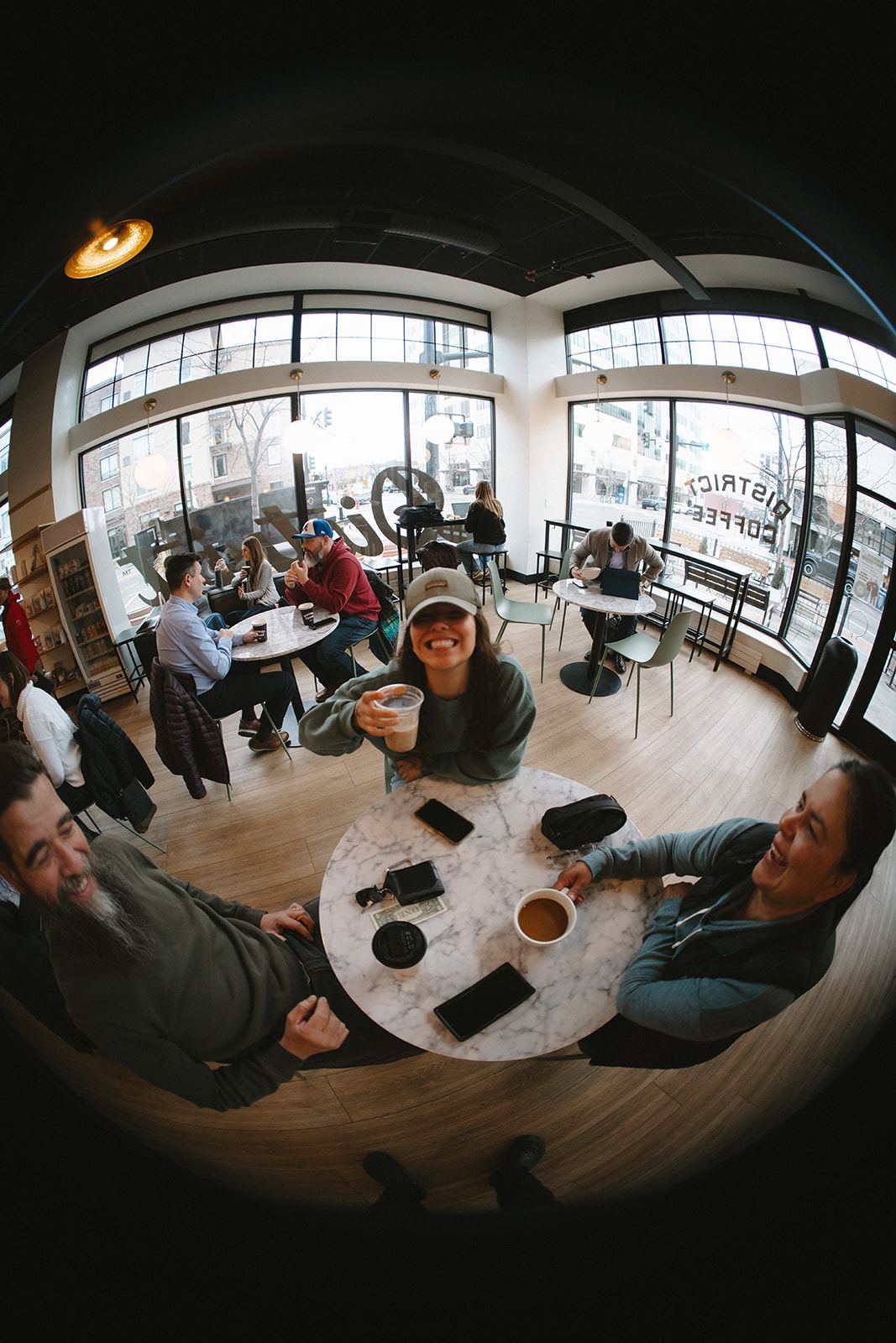 Fisheye view of the busy District Coffee House with a customer in a cap raising her coffee and two friends laughing at a marble table