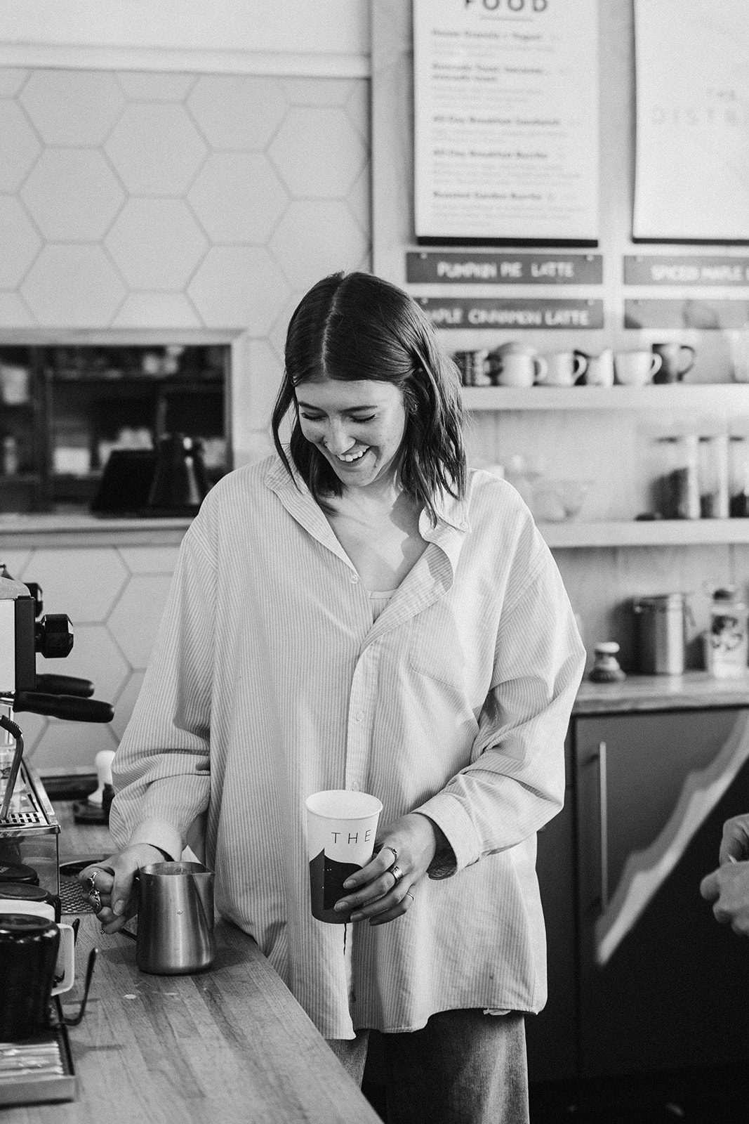 Black and white photo of a barista in an oversized striped shirt laughing as she looks down at a pitcher and cup behind the bar