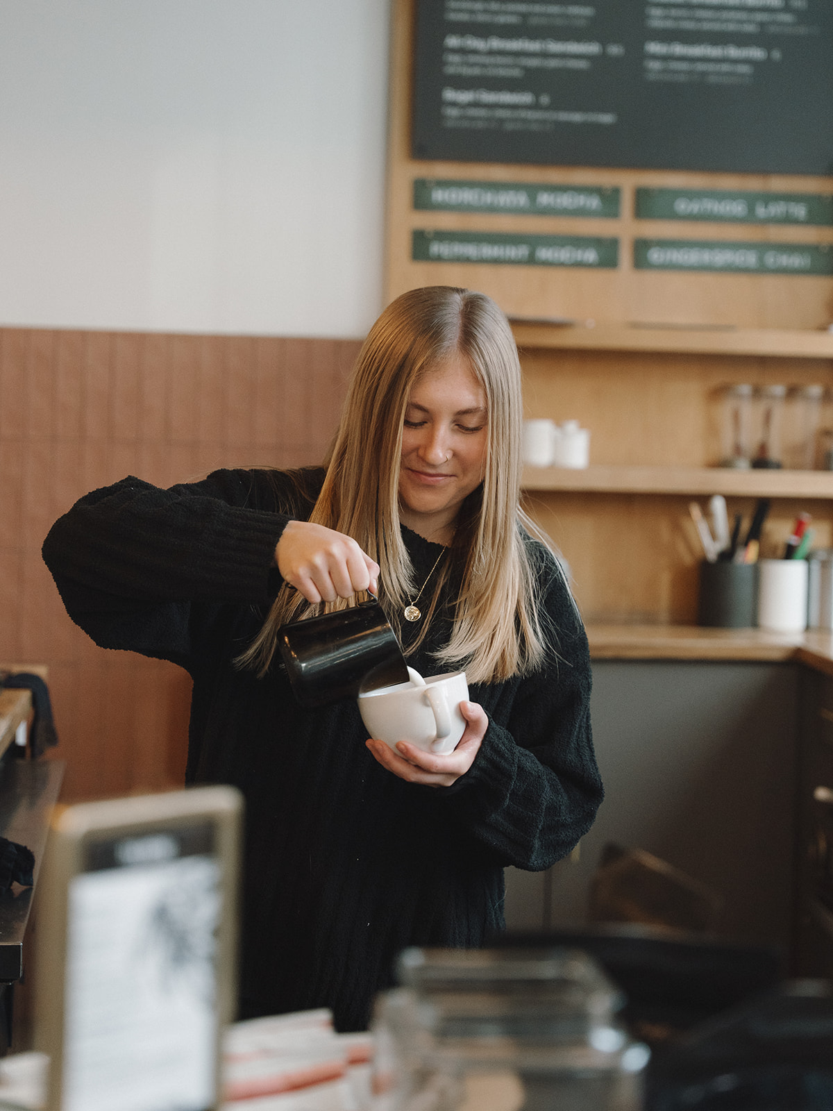 Barista with long blonde hair smiling down as she pours steamed milk into a white cup at the District Coffee House bar, with the menu board behind her