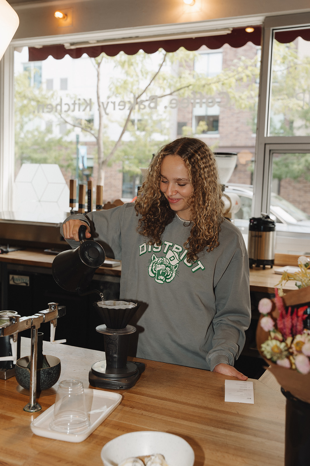 Barista in a gray District sweatshirt with curly hair pouring hot water from a kettle over a pour-over coffee