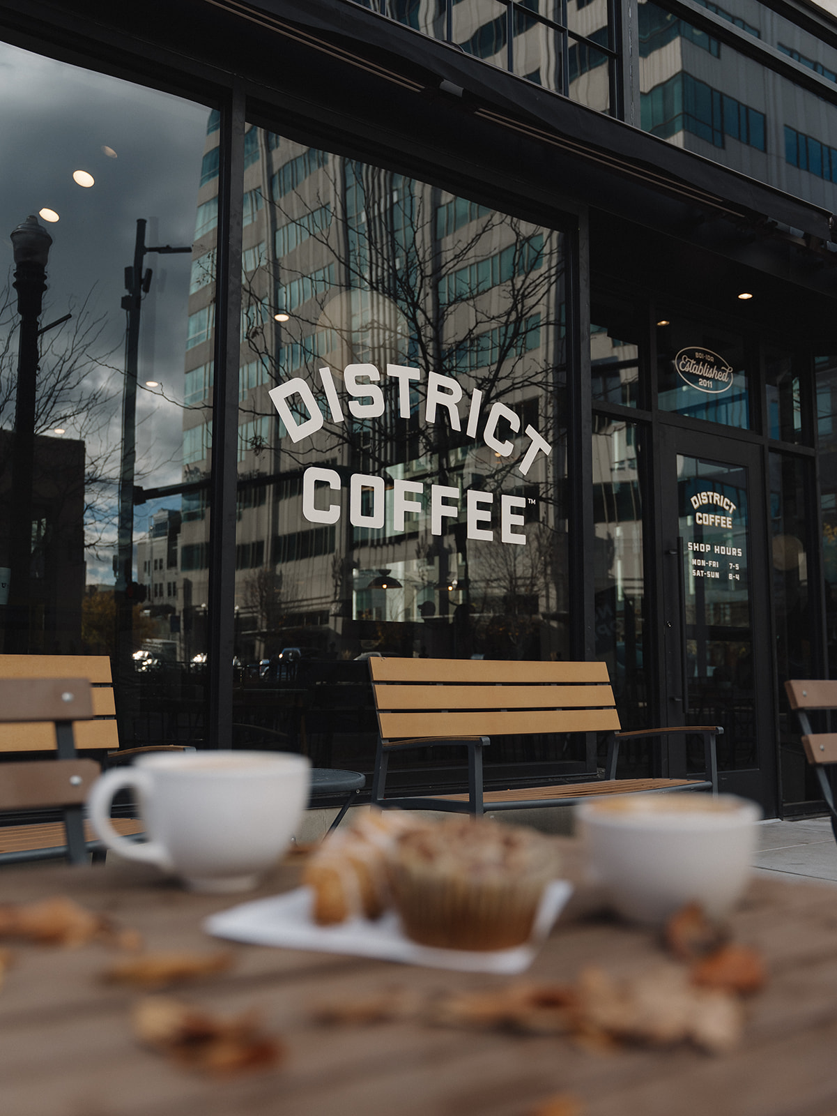 District Coffee window lettering reflecting downtown buildings, with two blurred lattes and a pastry on a patio table in the foreground