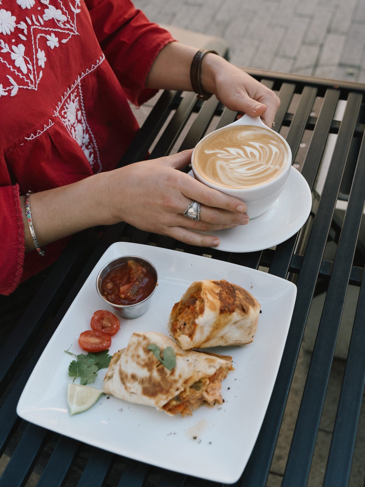 Hands holding a latte with a leaf pattern next to a breakfast burrito cut in half with salsa and lime