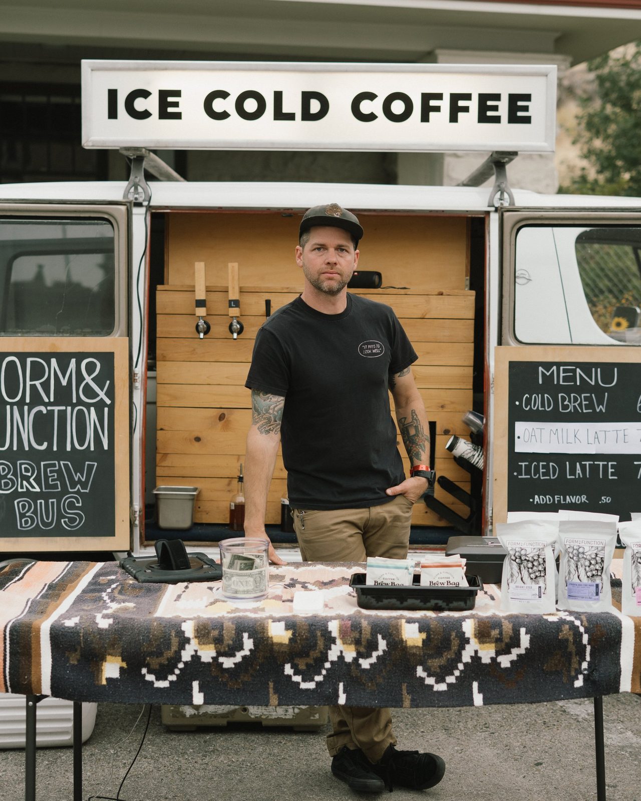 Man standing behind a table at the Form & Function Brew Bus, a van with an Ice Cold Coffee sign and chalkboard menus