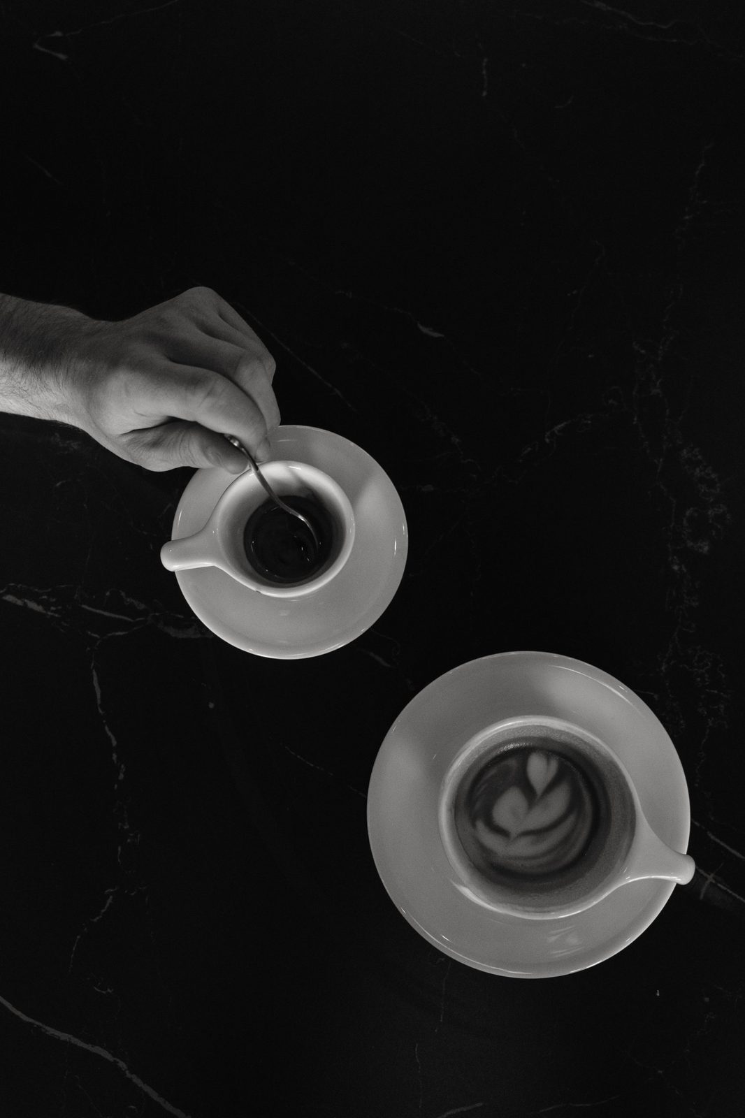 Black and white overhead of a hand stirring an espresso beside a latte on a dark marble table
