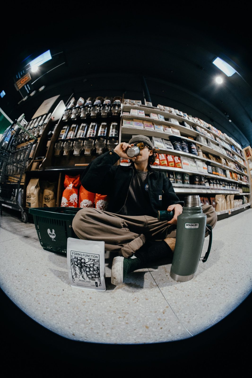 Fisheye photo of a man in a beanie drinking from a thermos cup, sitting cross-legged in a coffee aisle beside a bag of Form & Function coffee
