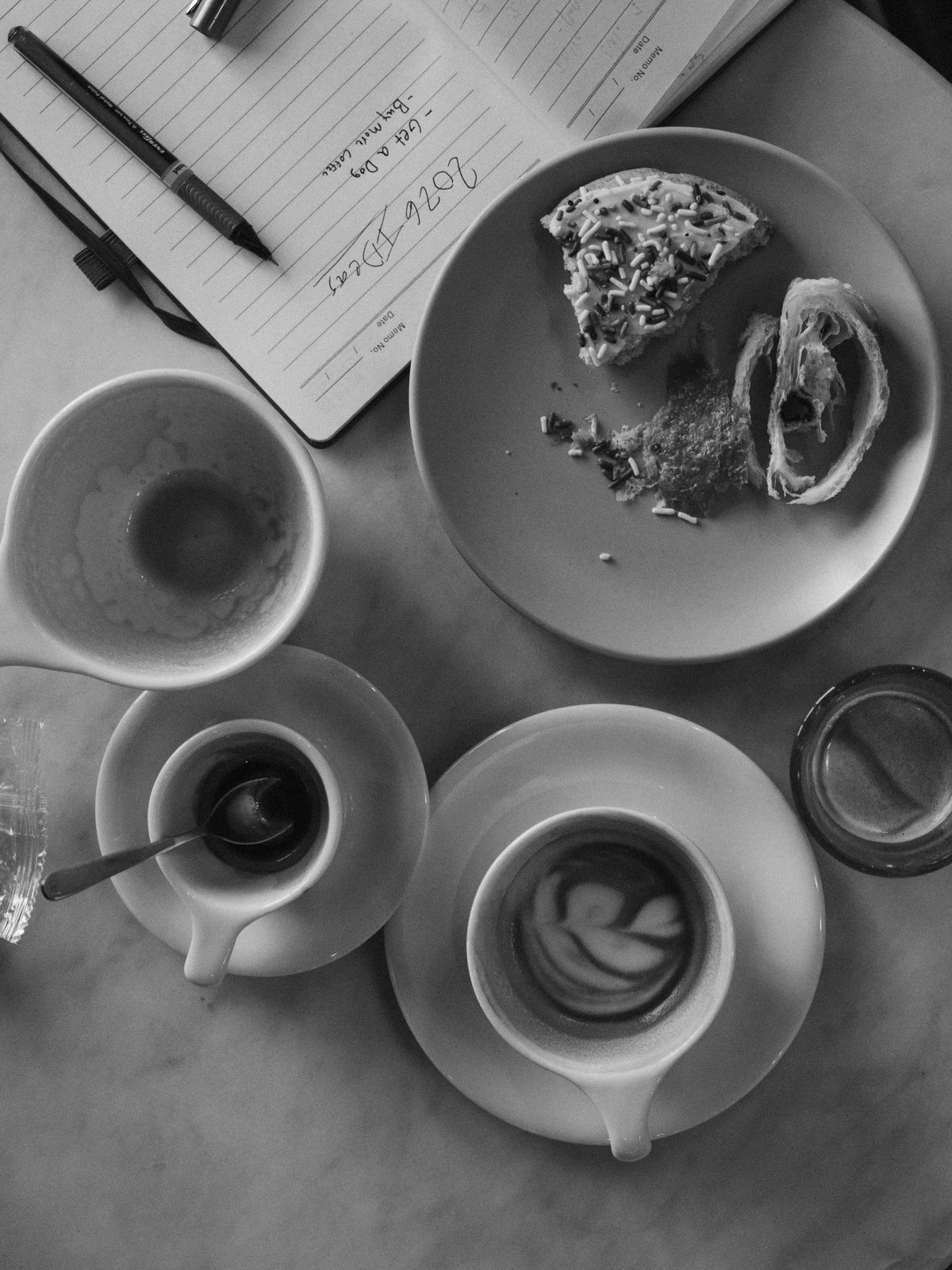 Black and white overhead of empty coffee cups, a half-eaten pastry and an open notebook on a marble table