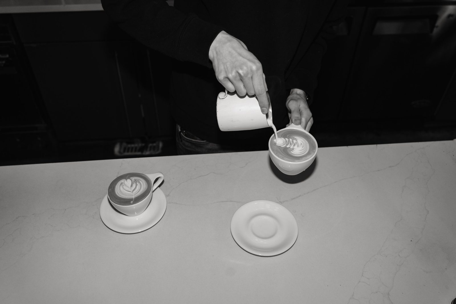 Black and white shot of a barista pouring milk into a latte, with a finished latte and an empty saucer on the counter