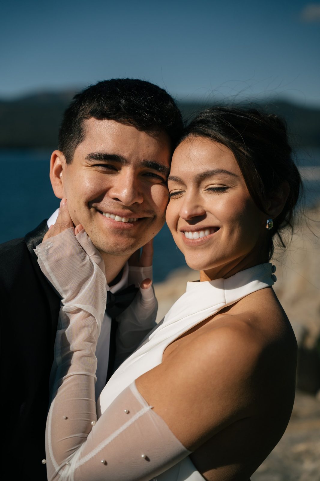 Smiling couple cheek to cheek in the sun, the bride in a white halter dress with sheer pearl sleeves, a lake behind them