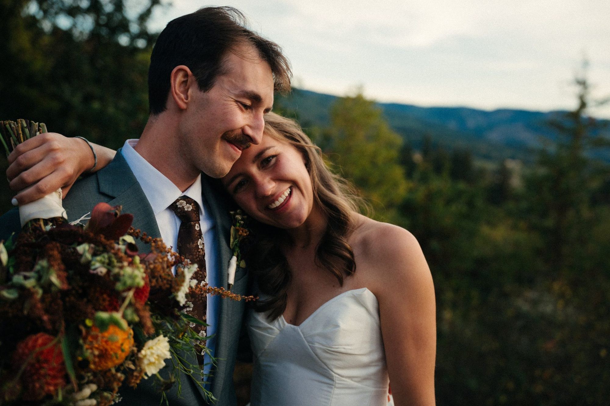 Groom smiling with his cheek against his bride's head as she laughs, holding an autumn bouquet with mountains behind them
