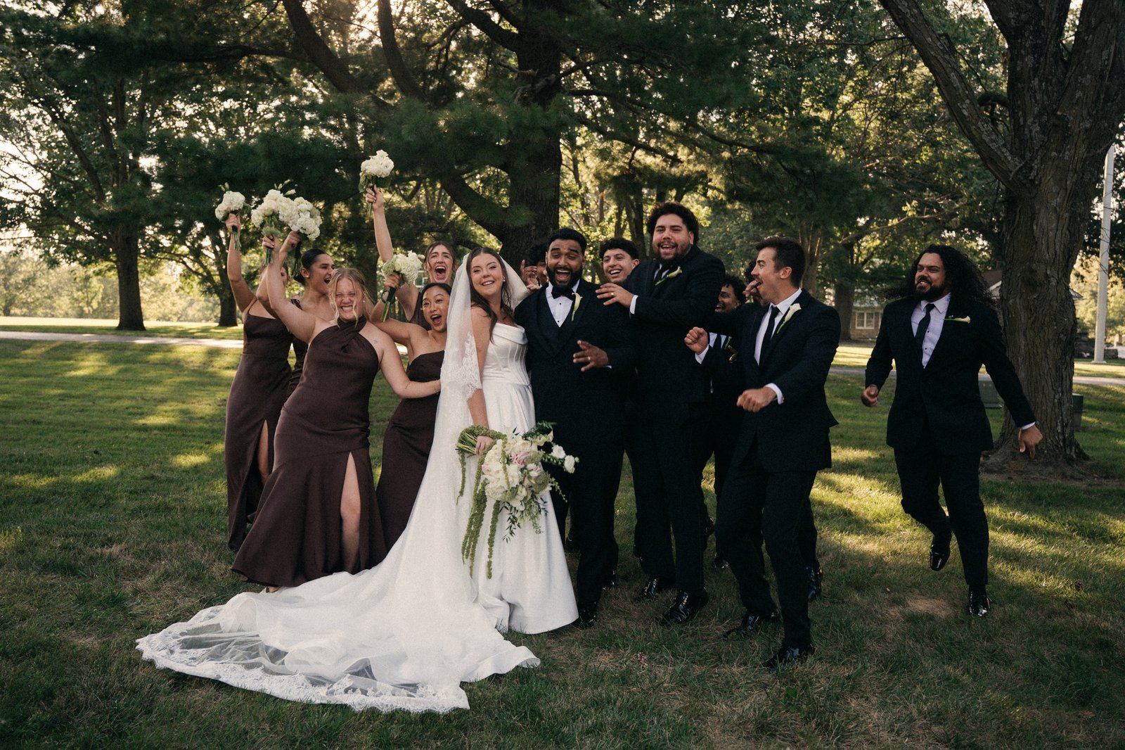A bride and groom surrounded by cheering bridesmaids holding white bouquets and groomsmen jumping in and laughing under tall trees