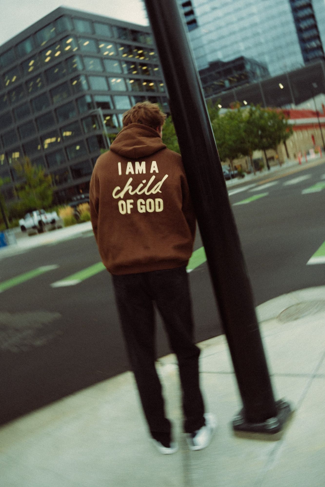 Person seen from behind in a brown I am a child of God hoodie, leaning on a street pole with downtown buildings behind