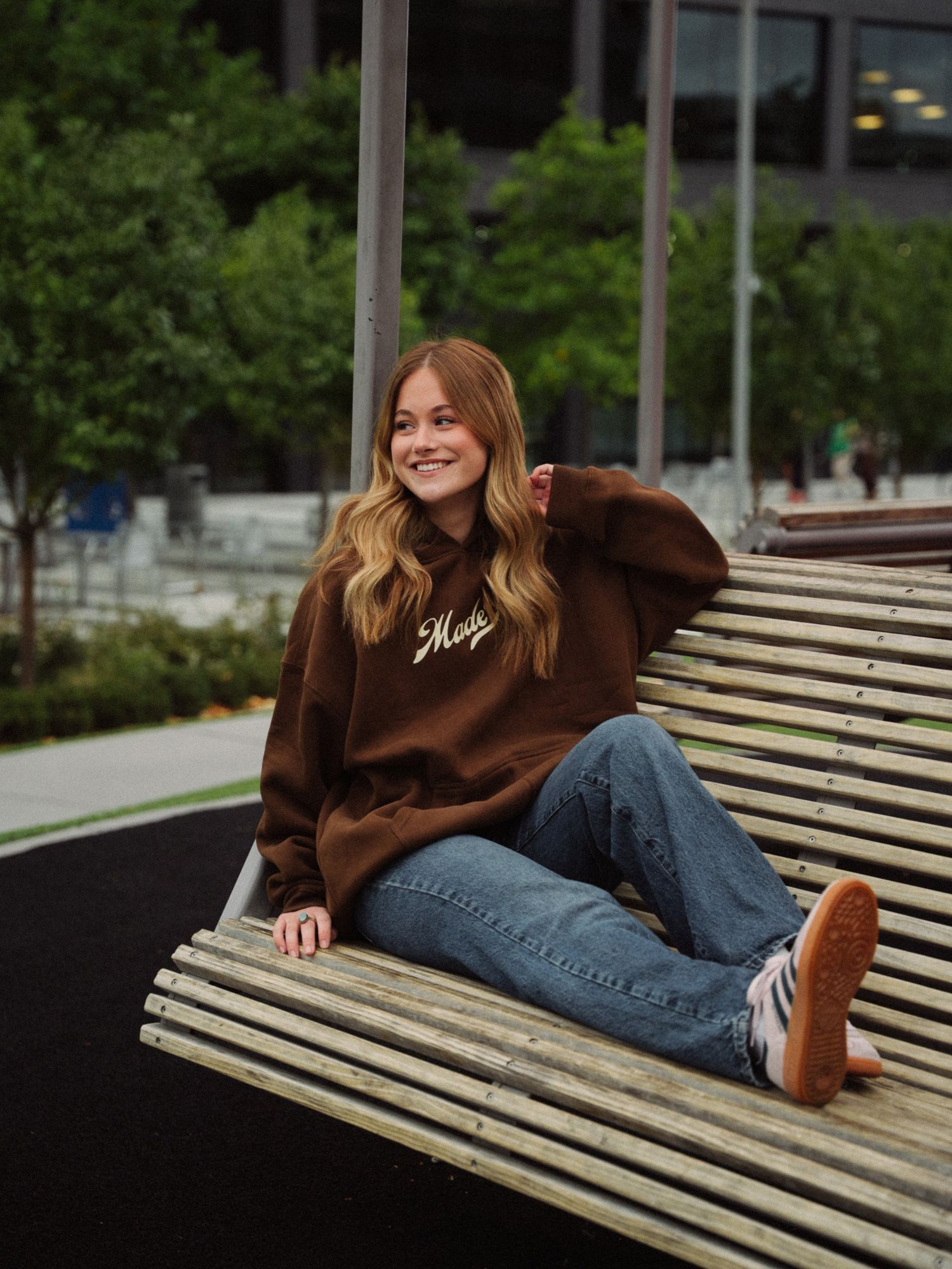 Woman in an oversized brown Made New hoodie and jeans, smiling and looking off to the side from a slatted wooden bench
