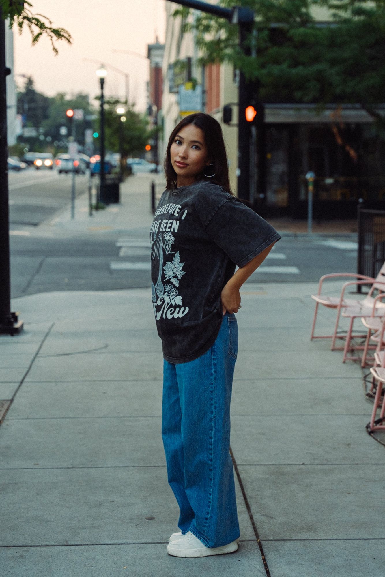 Woman standing in profile on a downtown sidewalk with a hand on her hip, looking back at the camera in a washed black graphic tee