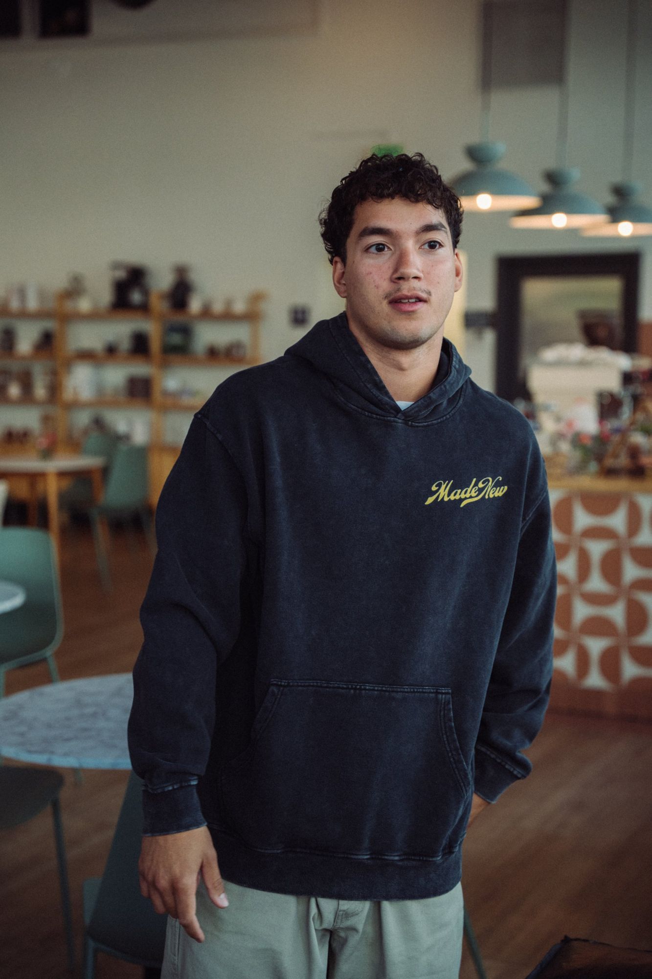 Man in a washed black Made New hoodie with a yellow script logo, standing in a bright cafe with green chairs and wooden shelves