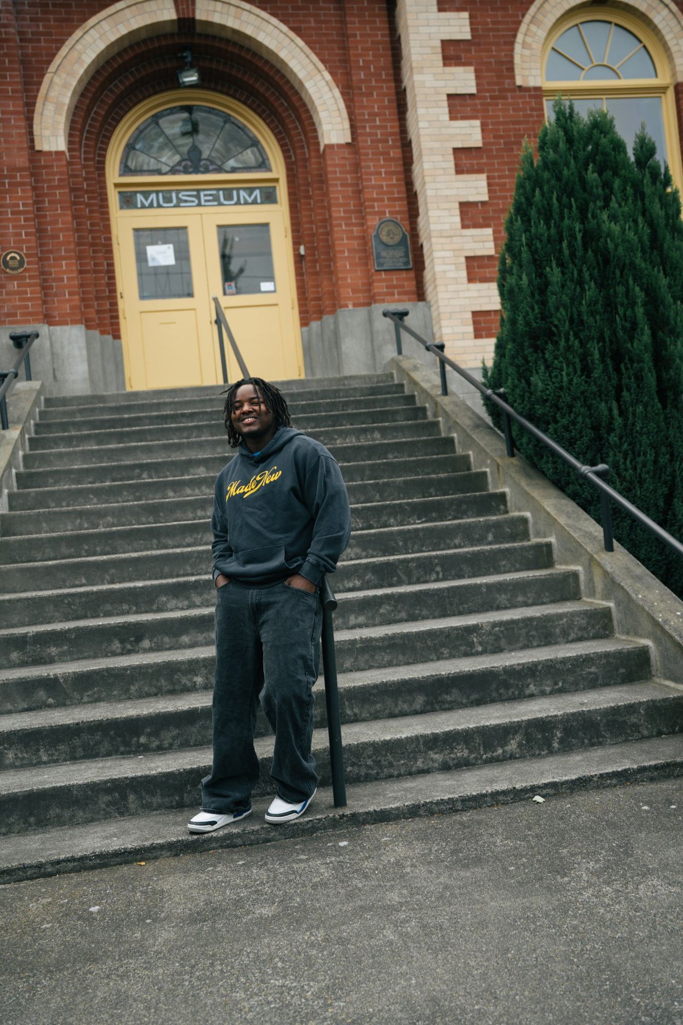Smiling man in a washed Made New hoodie with a yellow logo, leaning on a railing on the steps of a brick museum building