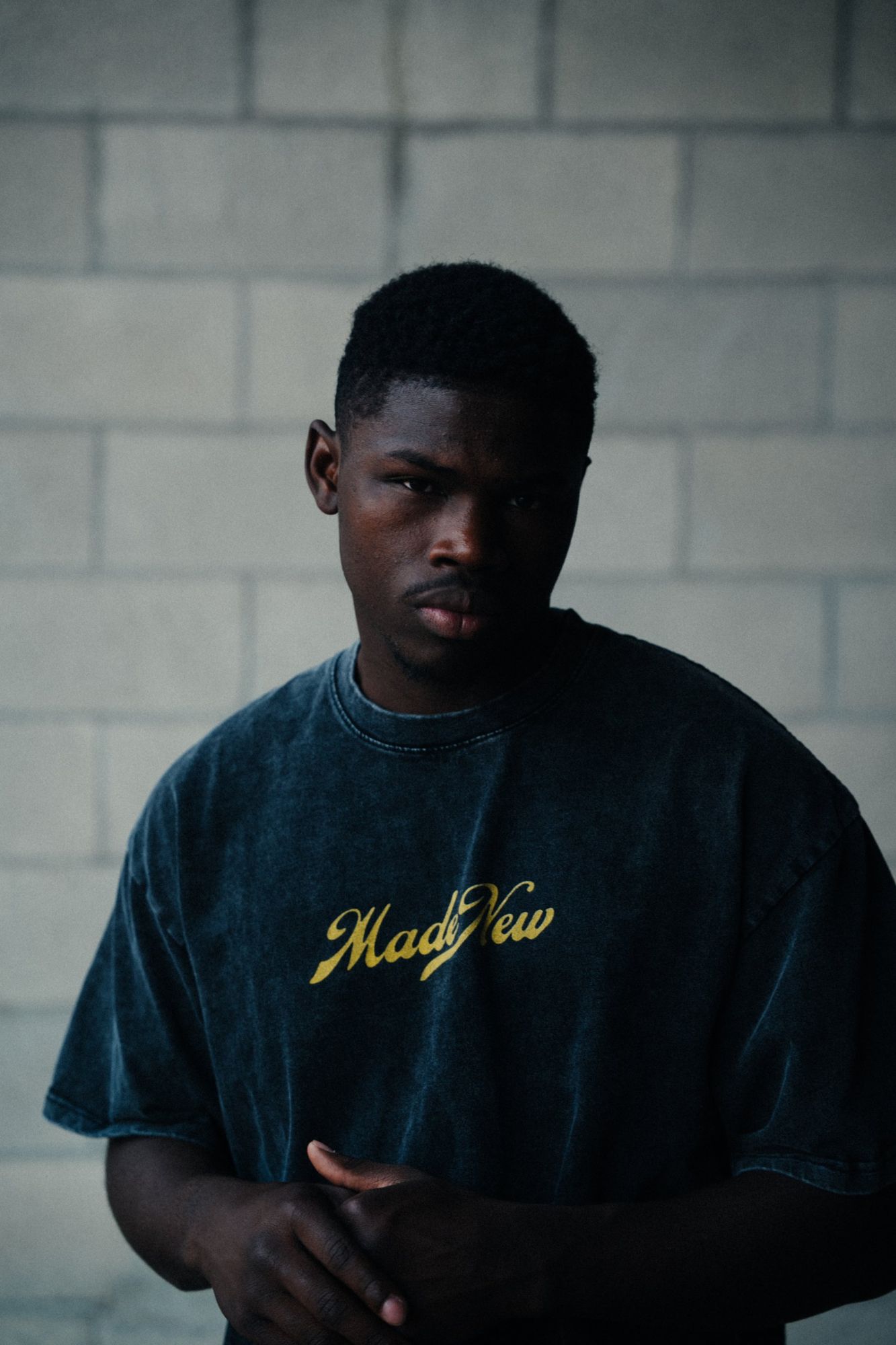 Serious portrait of a man in a washed navy Made New tee with a yellow script logo, in front of a cinder block wall