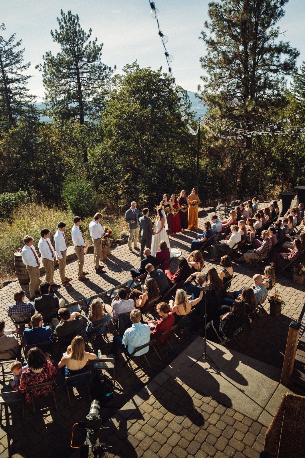 Overhead view of an outdoor wedding ceremony on a stone patio with guests in folding chairs, groomsmen lined up and pine trees behind