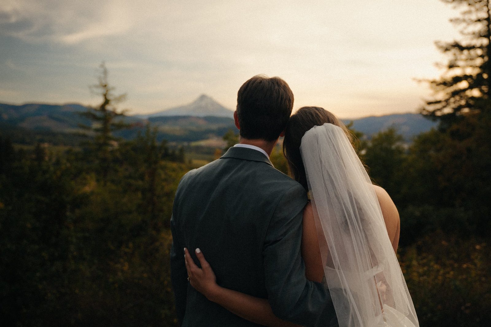 Bride rests her head on the groom's shoulder as they look out at Mount Hood at sunset, her veil hanging down her back