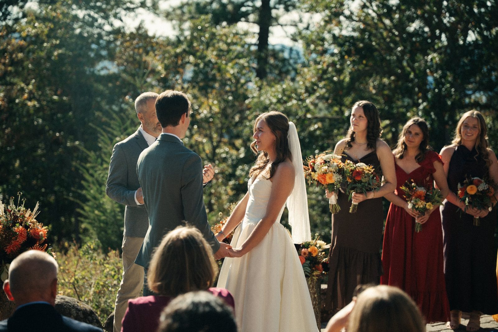 Bride and groom hold hands facing each other during their ceremony at the Crag Rat Hut in Hood River, Oregon, bridesmaids in autumn dresses behind them