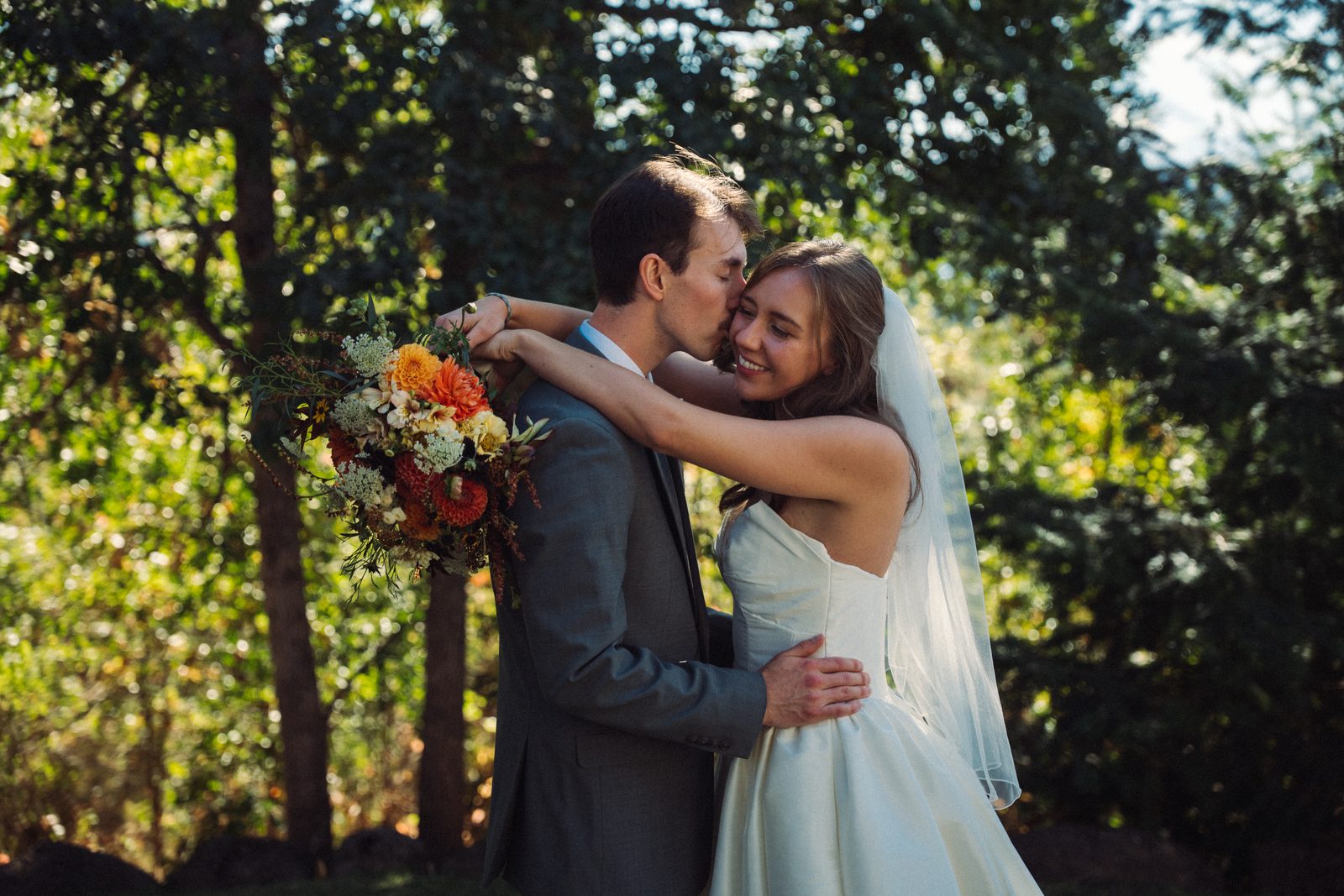 Groom kisses his bride on the cheek as she wraps her arms around him, bouquet over his shoulder