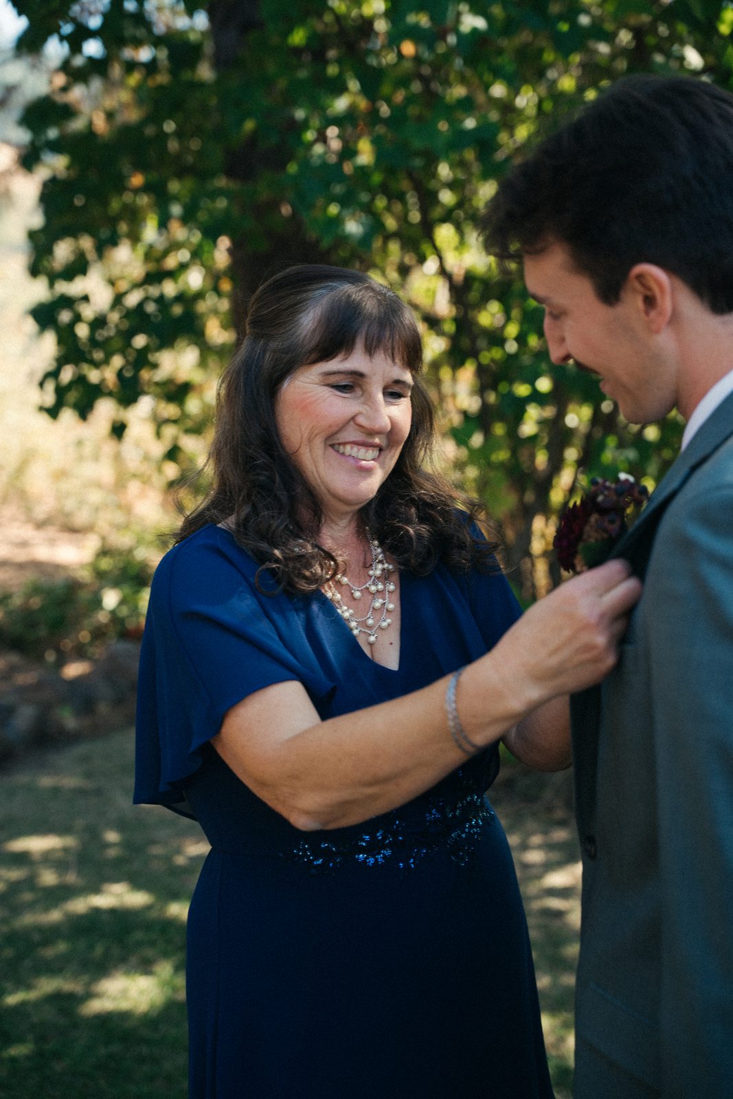 Groom's mother in a navy dress smiling as she pins his boutonniere